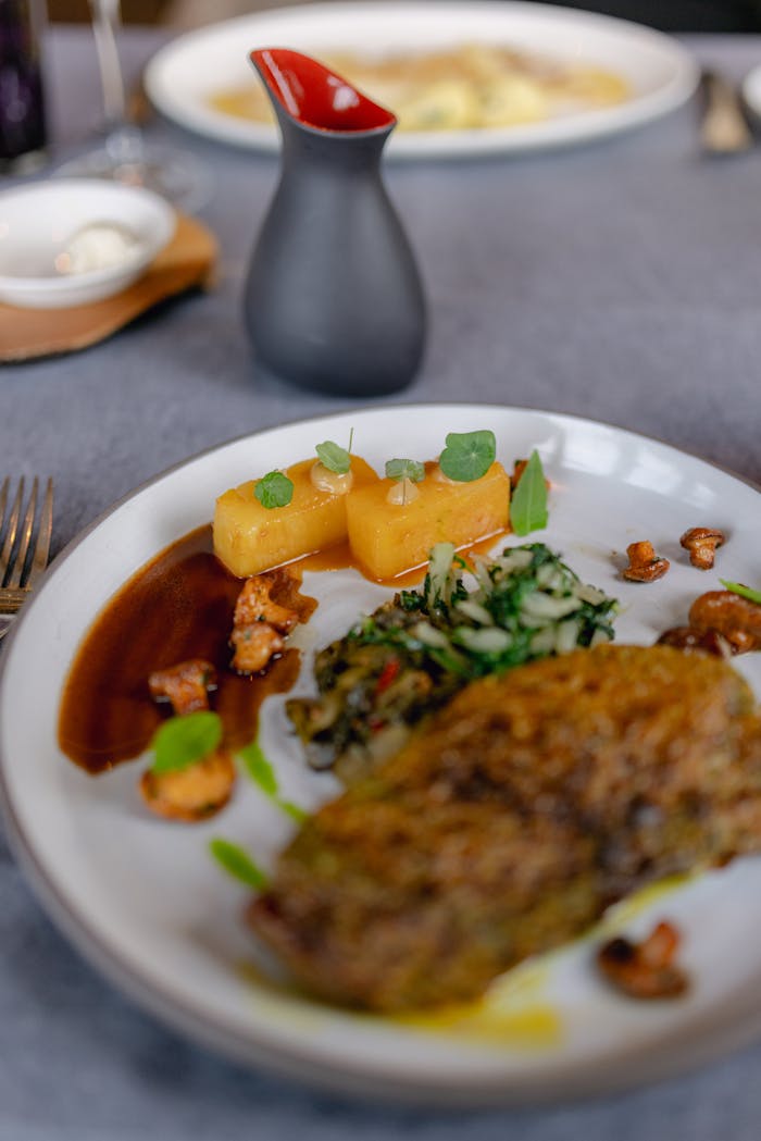 A gourmet dish featuring a main meat portion, potatoes, greens, and a dark sauce, elegantly plated.